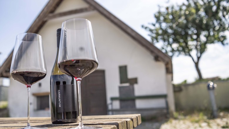 Zwei Weingläser und eine Flasche auf Holztisch vor kleinem Gebäude., © Donau Niederösterreich, Robert Herbst