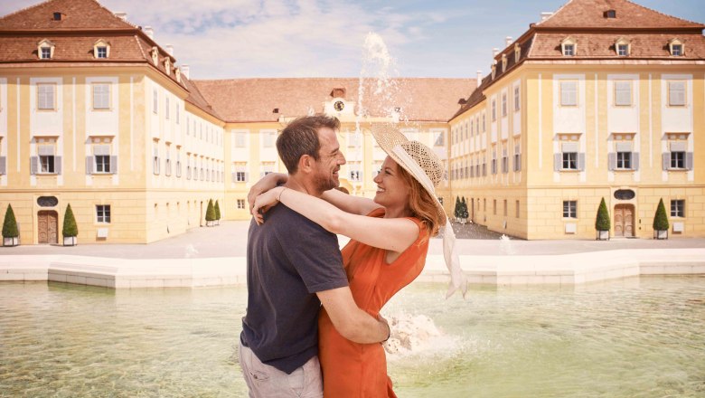 Paar umarmt sich vor einem barocken Schloss mit Brunnen., © Donau Niederösterreich Tourismus, Andreas Hofer