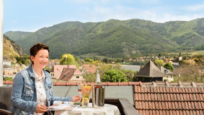 Frau genießt Wein auf Terrasse mit Blick auf grüne Hügel und Dorf., © DI Doris Hamberger