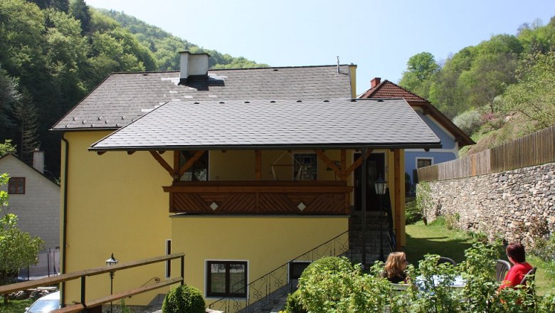 Gelbes Haus mit Balkon, umgeben von grüner Landschaft und Steinmauer., © Bergkirchner